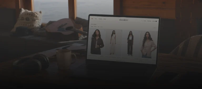 Notebook em uma mesa de escritório exibindo a página de e-commerce da Shoulder, com xícaras, fones e artigos de trabalho ao fundo em ambiente interno.